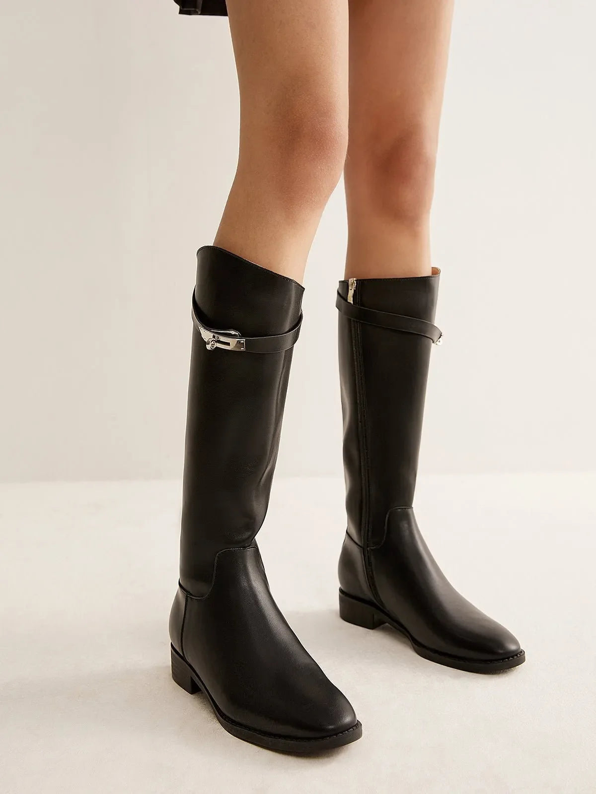 Pair of black knee-high buckle boots worn with bare legs, showing the front and side zipper.