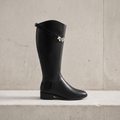 Black knee-high riding-style boot with a low heel and silver-tone buckle against a light stone backdrop.